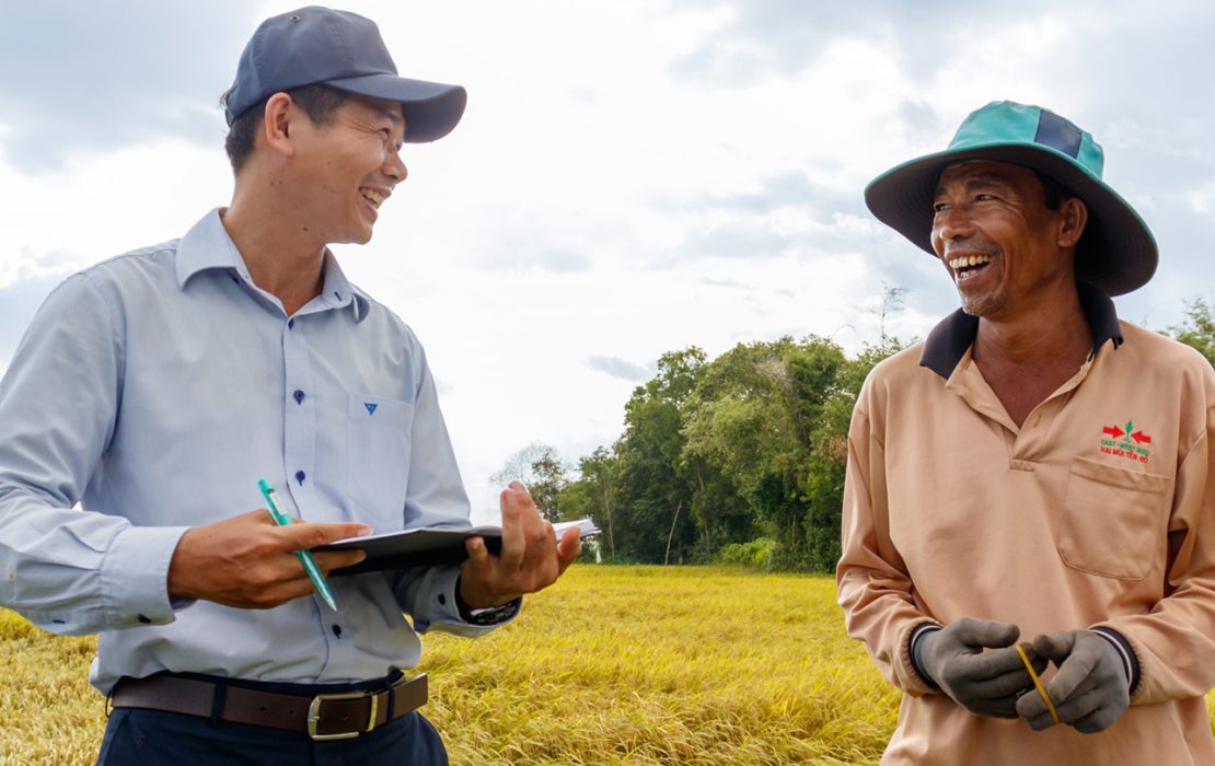 Un agricultor vietnamita sonriente y un funcionario del proyecto conversan en un campo de arroz, destacando los esfuerzos de adaptación climática impulsados ​​por la comunidad