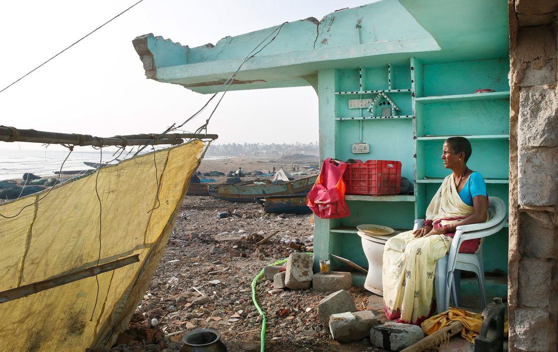 Woman sits inside a damaged coastal structure in Odisha, India, after cyclones and flooding, with debris and fishing boats visible along the shore.