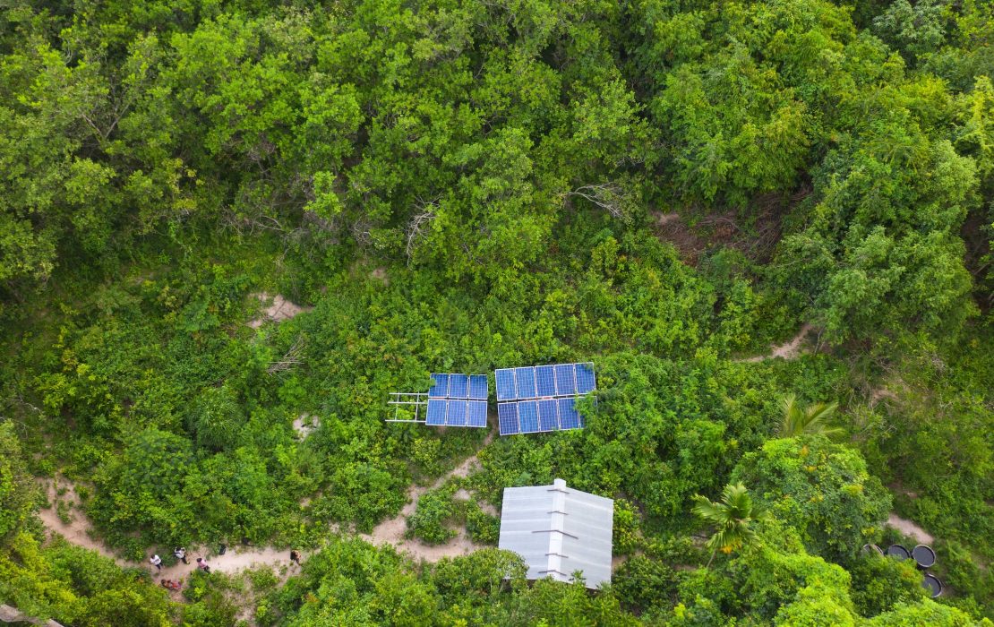 Aerial view of a small solar-panel installation and a metal-roofed structure surrounded by dense green forest.