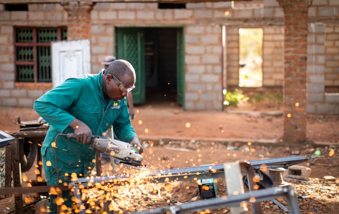 21 African countries will achieve economic growth exceeding 5 percent in 2025. Photo: UNDP Zimbabwe A man in a work uniform and safety goggles uses a power grinder on metal, producing bright sparks, at a construction site with an unfinished brick building in the background in Zimbabwe. Photo: UNDP Zimbabwe