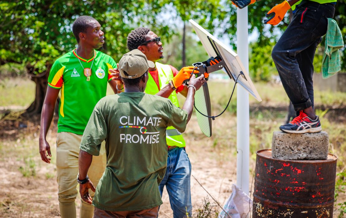 Renewable energy solutions like solar-powered street lighting help empower local communities and create green jobs. Photo: UNDP Senegal A technician in a high-visibility vest and orange gloves installing a solar-powered LED street light on a tall pole, with trees and a clear blue sky in the background in Casamance, Senegal