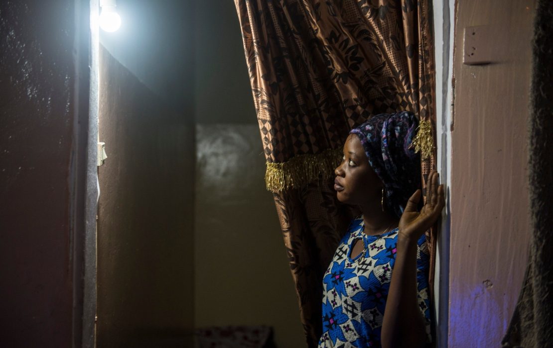 While Senegal has one of the highest electrification rates in West Africa, significant disparities remain among households. Photo: UNDP Senegal A woman in Senegal wearing a patterned blue dress and headscarf stands in a doorway, gently holding back a curtain. A single lightbulb glows above her, casting soft shadows on the wall.