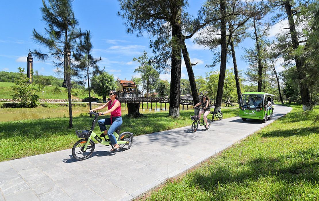 Women cycling using e-bikes in a park in Hue, followed by an electric bus under a blue sky.