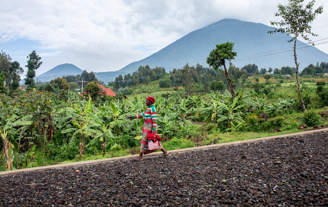 Woman walks along a rural road in Burera District, Rwanda, with banana fields and forested hills in the background.