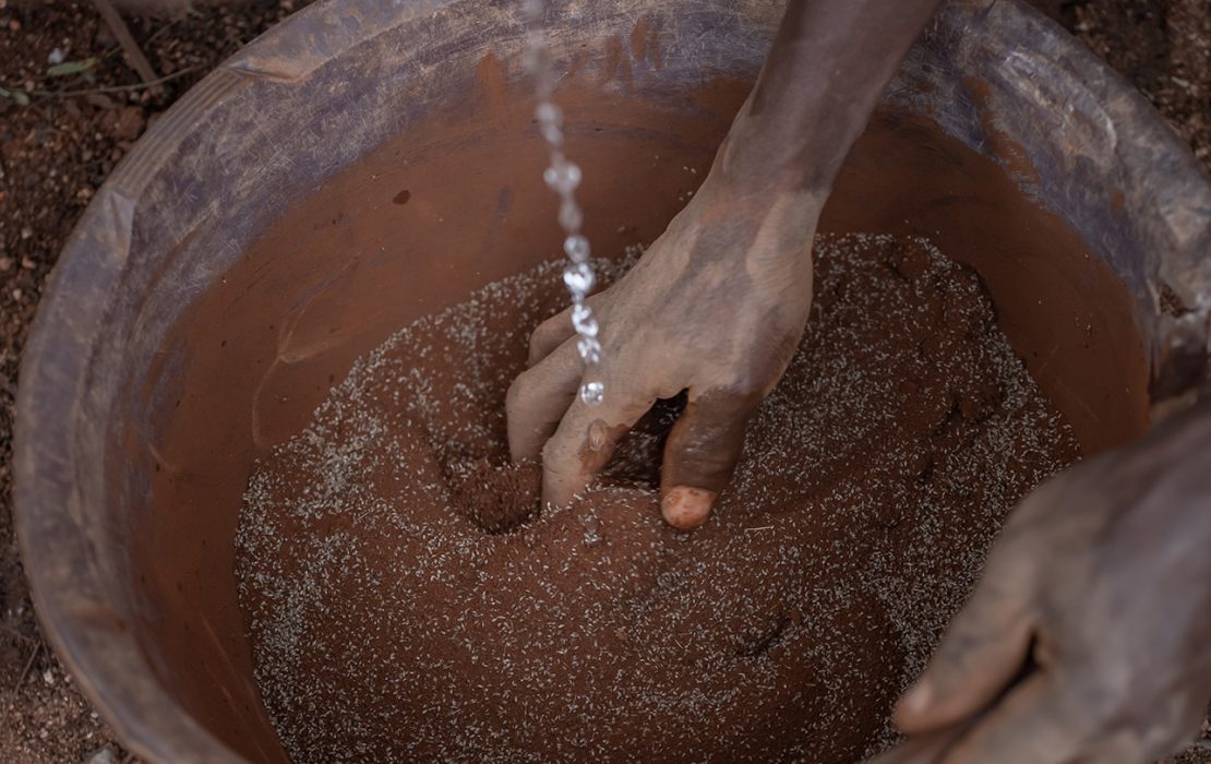  A person’s hand mixes seeds with soil in a clay pot as water is poured in, preparing the seeds for planting in a community-led conservation effort in Tanzania.