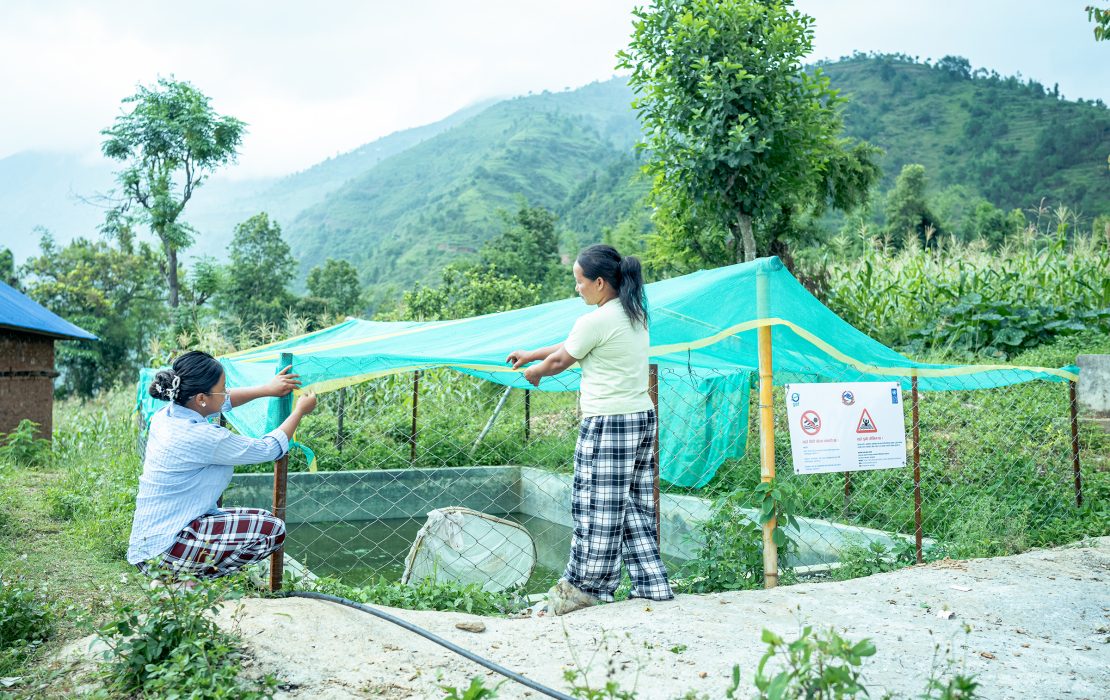 Two women secure a green protective net over a fenced water storage pond in a rural hillside area, with terraced fields and mountains in the background. 
