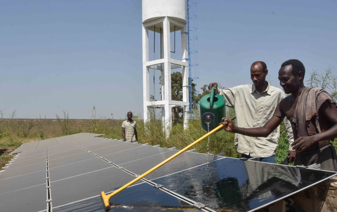 Limited access to equipment and lack of financing are major challenges hindering broader adoption of solar energy in Senegal. Photo: UNDP Senegal Three Senegalese men work near a large solar panel array in an open field. Two are actively cleaning the panels using a watering can and a squeegee. A tall white water tower with a blue ladder stands in the background, surrounded by sparse vegetation.
