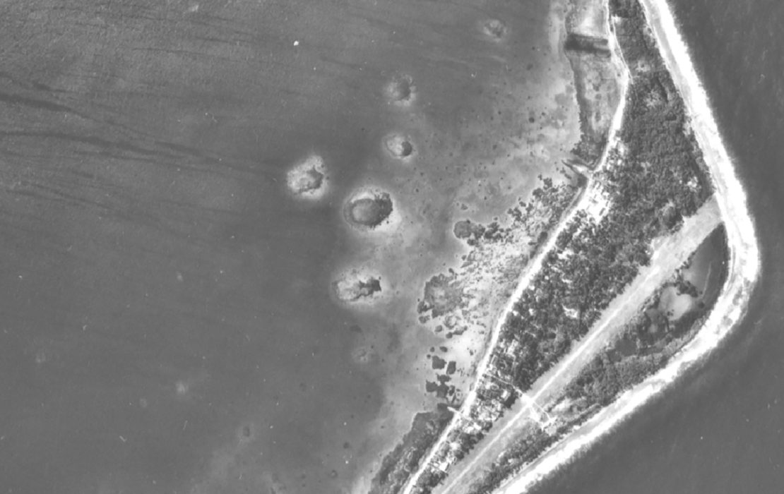 Black and white satellite image of atoll island showing lagoon and moonshaped thin strip of land with foliage and beaches.