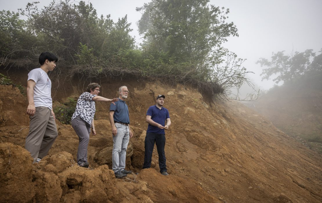 Forests are vital for Georgia’s climate goals and play a key role in preventing soil erosion. Photo: Irakli Dzneladze / UNDP Georgia In Georgia, four environmental field researchers stand on a steep, eroded hillside with exposed soil and sparse vegetation, analyzing landscape degradation. One person points toward the foggy horizon, indicating an area of concern. Trees and bushes line the top of the slope, suggesting a transitional ecosystem.
