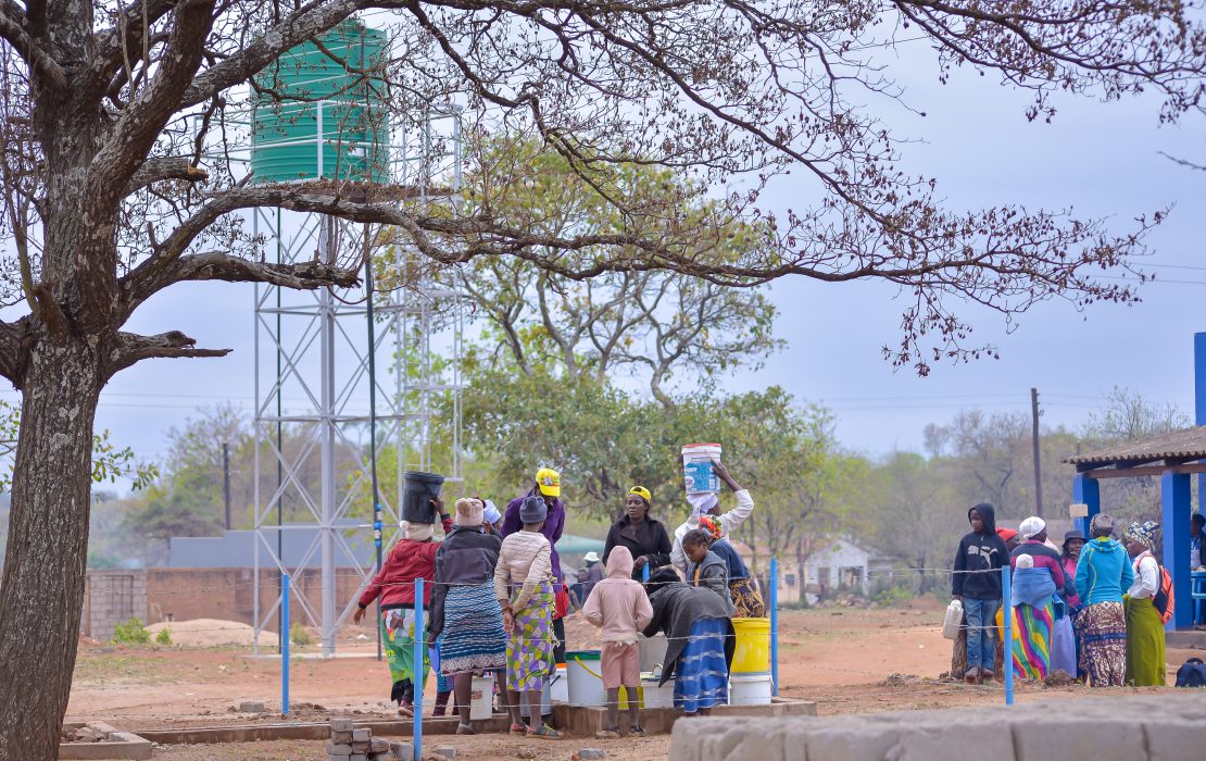 Renewable energy infrastructure provides access to water for productive and household use, including during droughts and floods. Photo: UNDP Zimbabwe Women in Zimbabwe gather around a rural water distribution point, collecting water from containers beneath a tall metal structure supporting a green tank.