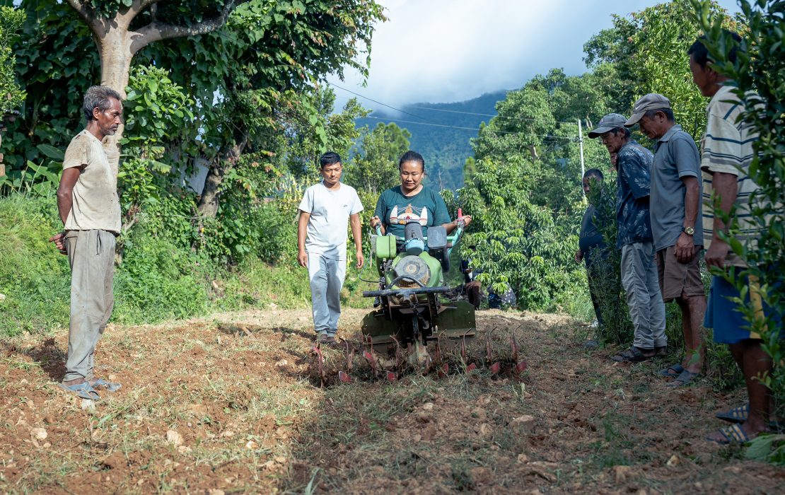 A group of farmers in rural Nepal watch as a woman uses a small power tiller to prepare the soil in a hillside field.