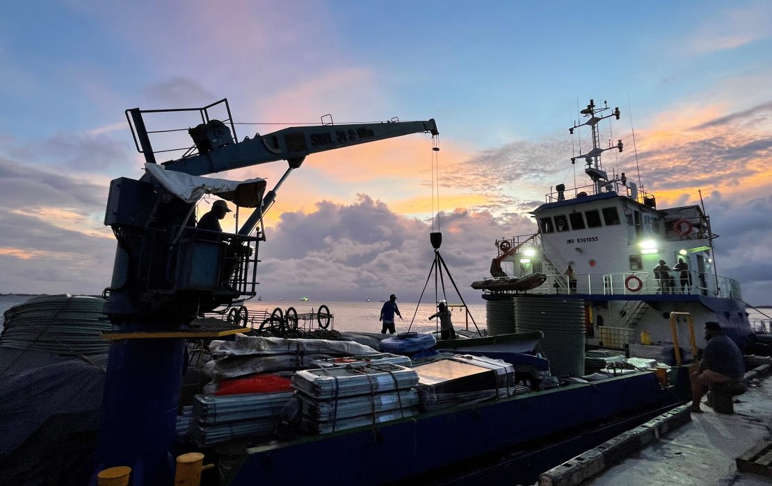 Photo: UNDP Pacific Office in Fiji Workers load supplies onto a docked vessel at sunset, with a crane lifting cargo against a colorful sky.