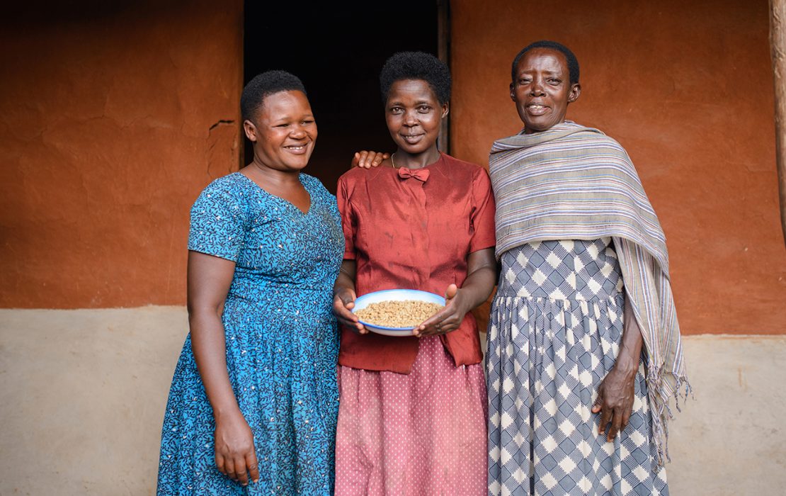 Three women standing together, smiling, with one holding a bowl of dried beans, symbolizing community collaboration and sustainable agricultural practices.