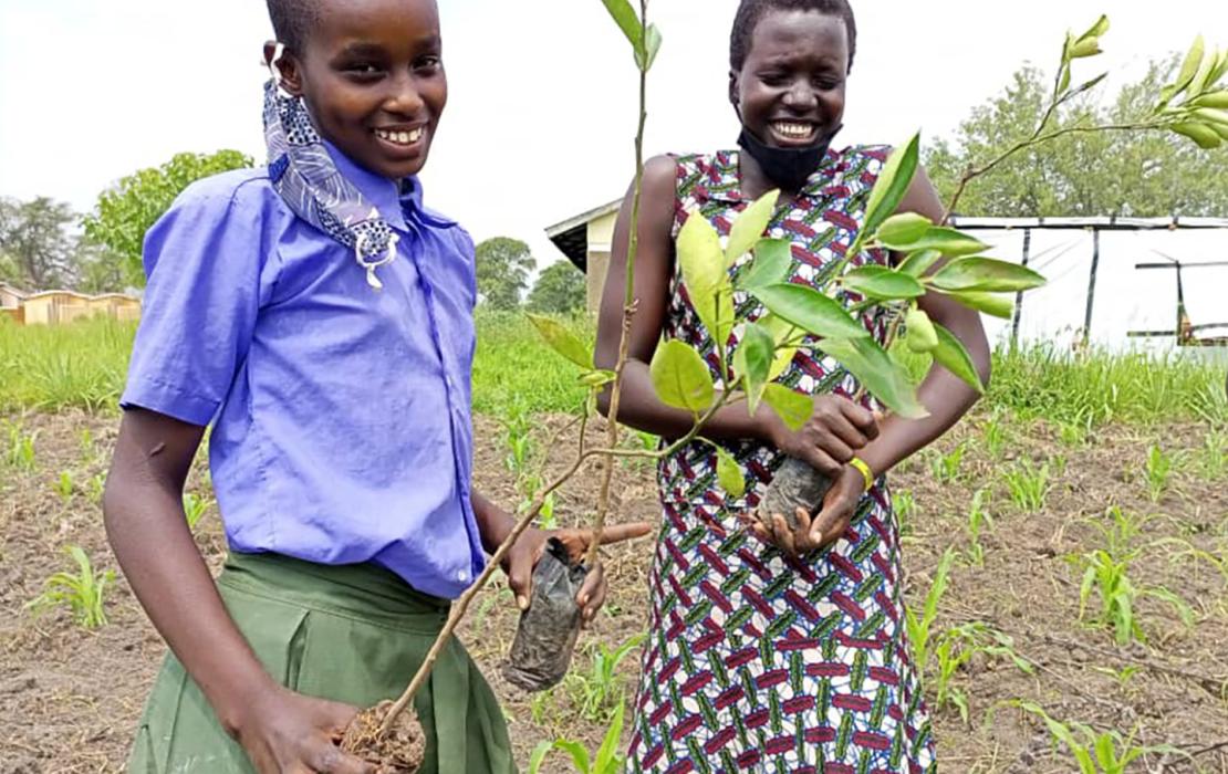 Two smiling young Ugandan women standing in a field, holding immature trees ready to plant. 