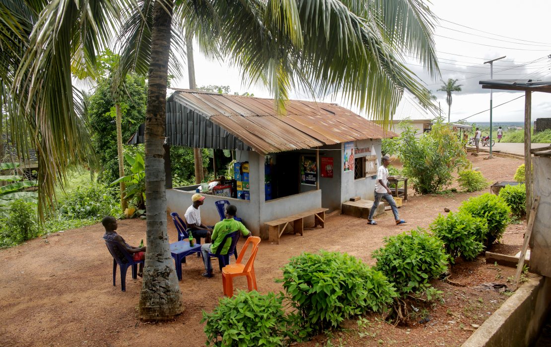 A small kiosk with a rusty roof under palm trees, with people sitting on plastic chairs in a leafy rural area.