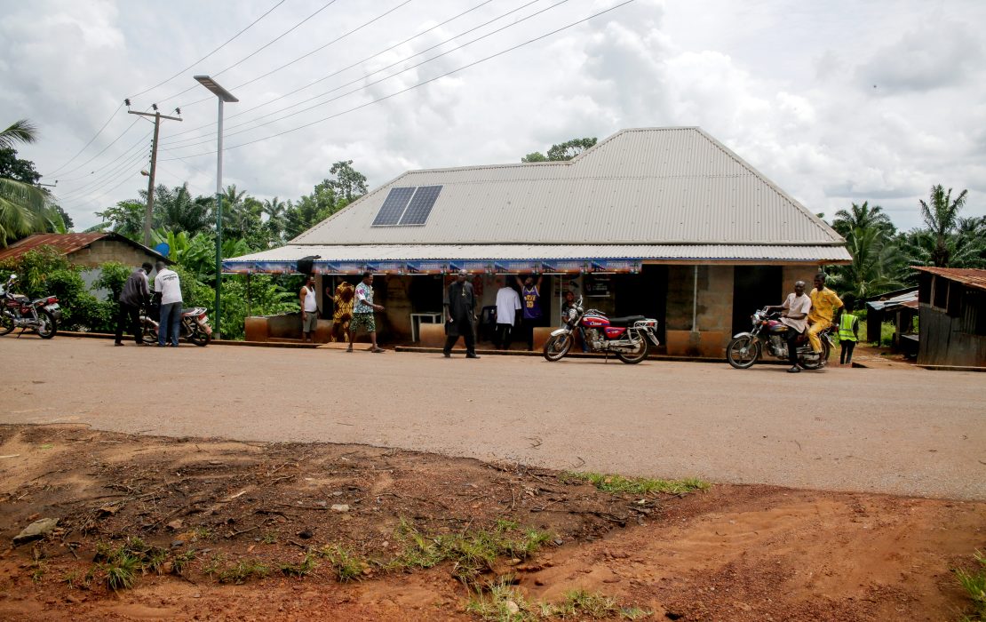 A roadside shop with a metal roof and a solar panel, with people and motorcycles gathered in a rural, green setting.