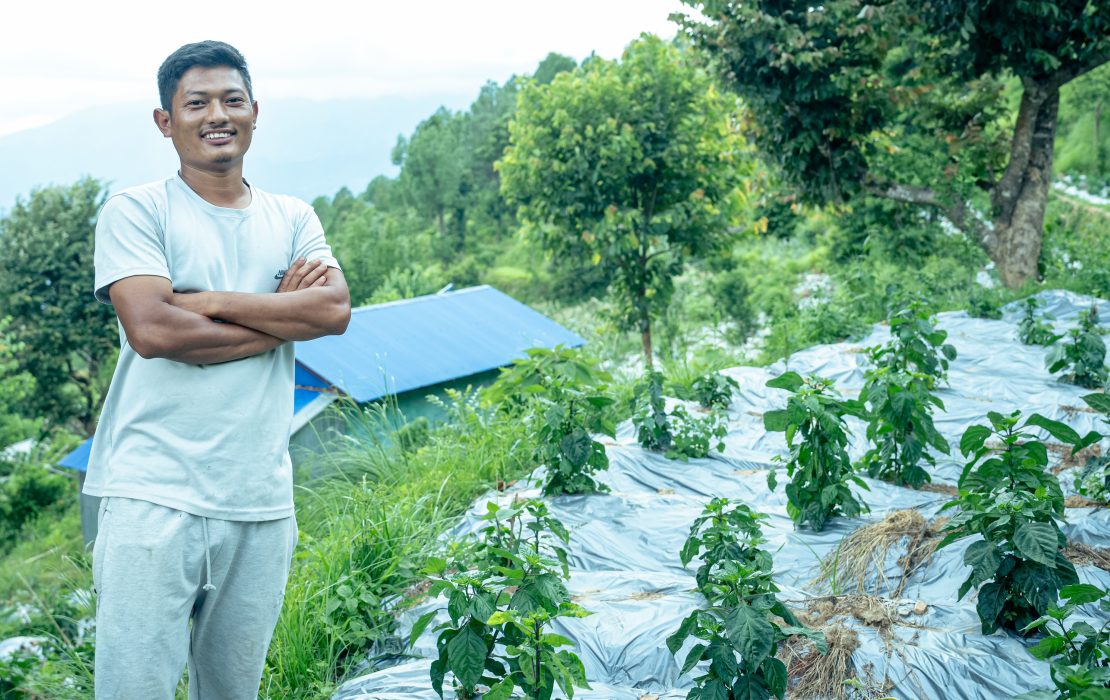 A smiling farmer stands with folded arms beside rows of young crops on terraced land, with green hills and a blue-roofed house in the background.