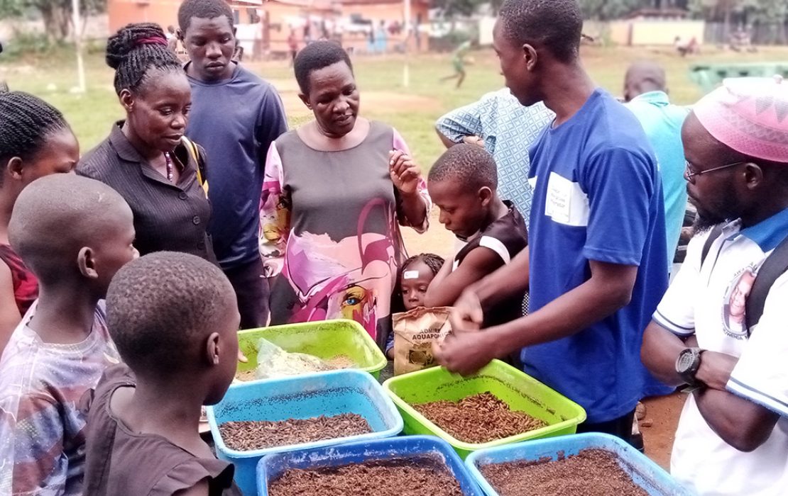 A group of community members, including children, gathers around trays of organic materials as a facilitator demonstrates sustainable farming practices during an outreach session in Uganda.