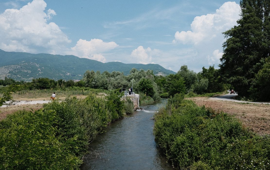 The diversion of the Sateska River to Lake Ohrid increased the risk of floods not just for the local communities along the river but also for the cities of Ohrid and Struga. Photo: Ariel Schindler/Adaptation Fund View of the Sateska River in North Macedonia flowing between green vegetation, with people standing on a small riverside structure and mountains in the background.