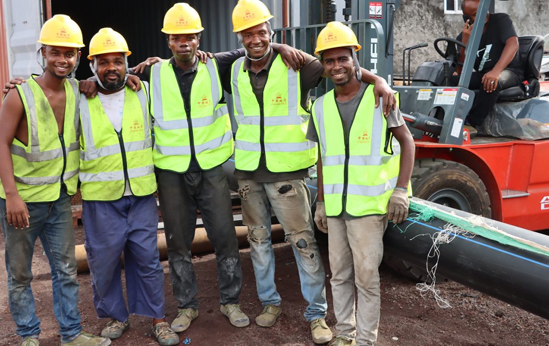 A group of construction workers in yellow hard hats and reflective vests smile while standing together at a worksite in Comoros