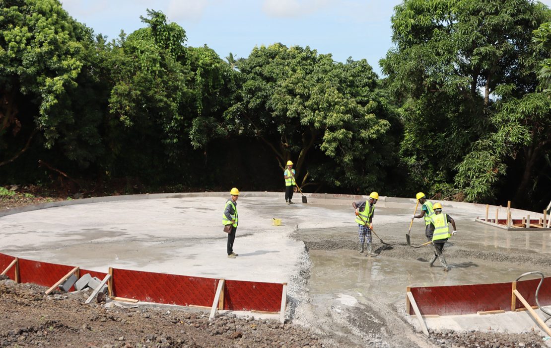 Construction workers wearing safety vests and helmets build a circular concrete water reservoir foundation surrounded by lush green trees in Comoros.