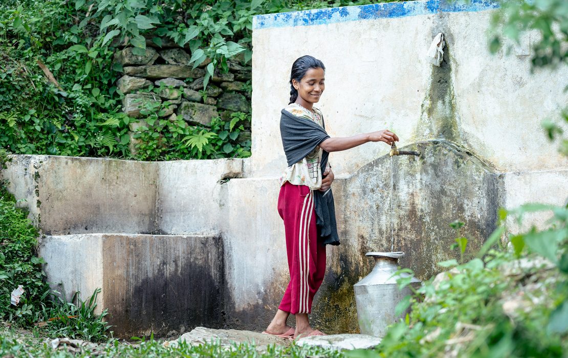 Woman collecting water from a community tap in rural Nepal, standing beside a water source surrounded by greenery.