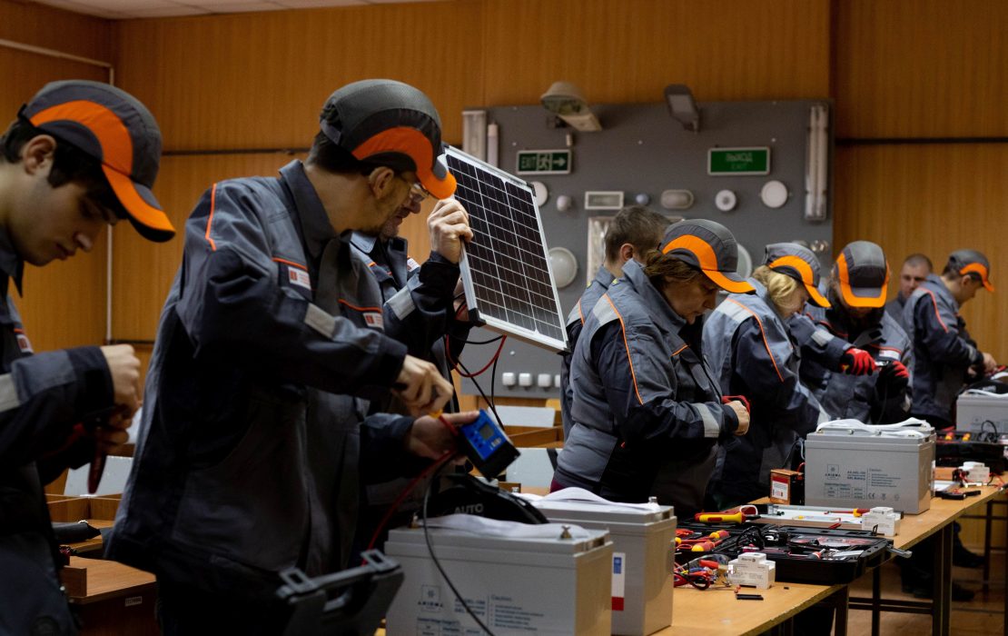 Electricians in Mykolaiv participate in a hands-on training course on renewable energy systems. Photo: UNDP Ukraine Group of workers in uniforms assembling and testing solar energy equipment indoors, including solar panels, batteries, and electrical tools in Ukraine