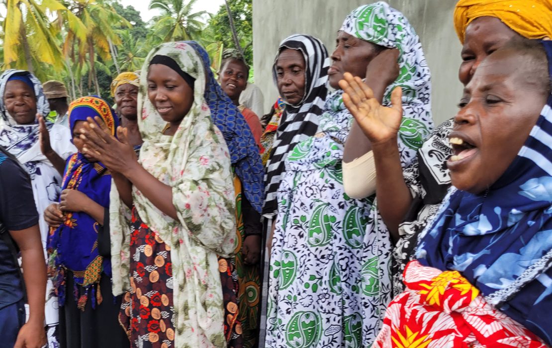 A group of women in colorful traditional clothing gathers outdoors, clapping and smiling as they celebrate newly installed water infrastructure.