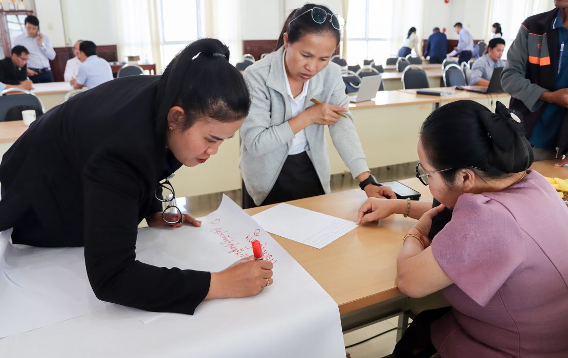 Three women collaborate around a table during a workshop, discussing ideas and writing notes on a large flip chart sheet.