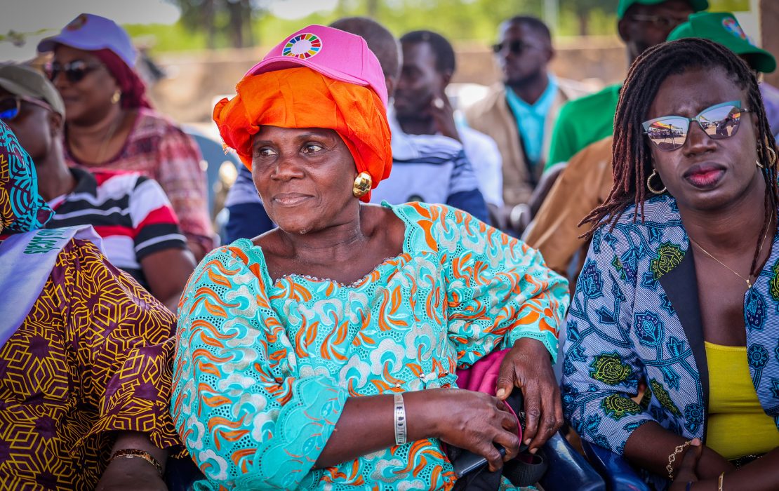 In Diogué island, access to renewable energy is changing the lives of women fish product processors and local communities. Photo: UNDP Senegal Women and community members in the island of Diogue in Casamance, Senegal, sit together at an outdoor event. A woman in a bright turquoise and orange dress with a matching headscarf and pink cap featuring the UN Sustainable Development Goals logo sits in the center, surrounded by others in colorful clothing.