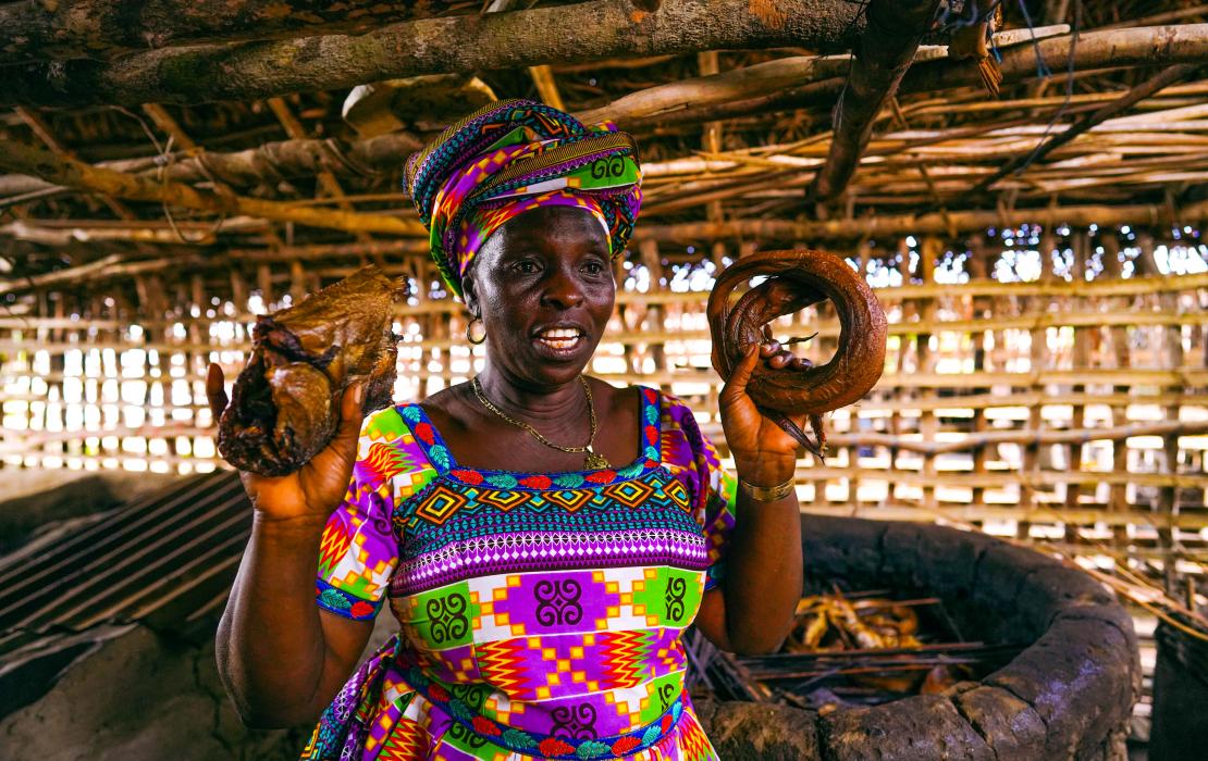 A fishmonger from Sierra Leone smiling and showing dried fishes in her local shop.