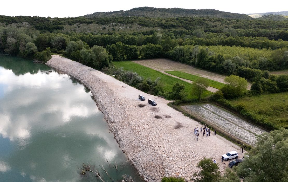 Aerial view of the restored Bojana/Buna River embankment in Montenegro, with cars and a small group of people gathered along the flood protection structure beside farmland and forest.