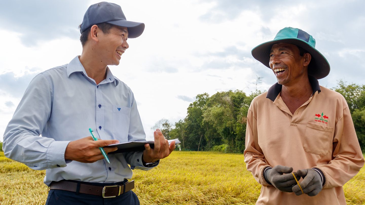 Un agricultor vietnamita sonriente y un funcionario del proyecto conversan en un campo de arroz, destacando los esfuerzos de adaptación climática impulsados ​​por la comunidad