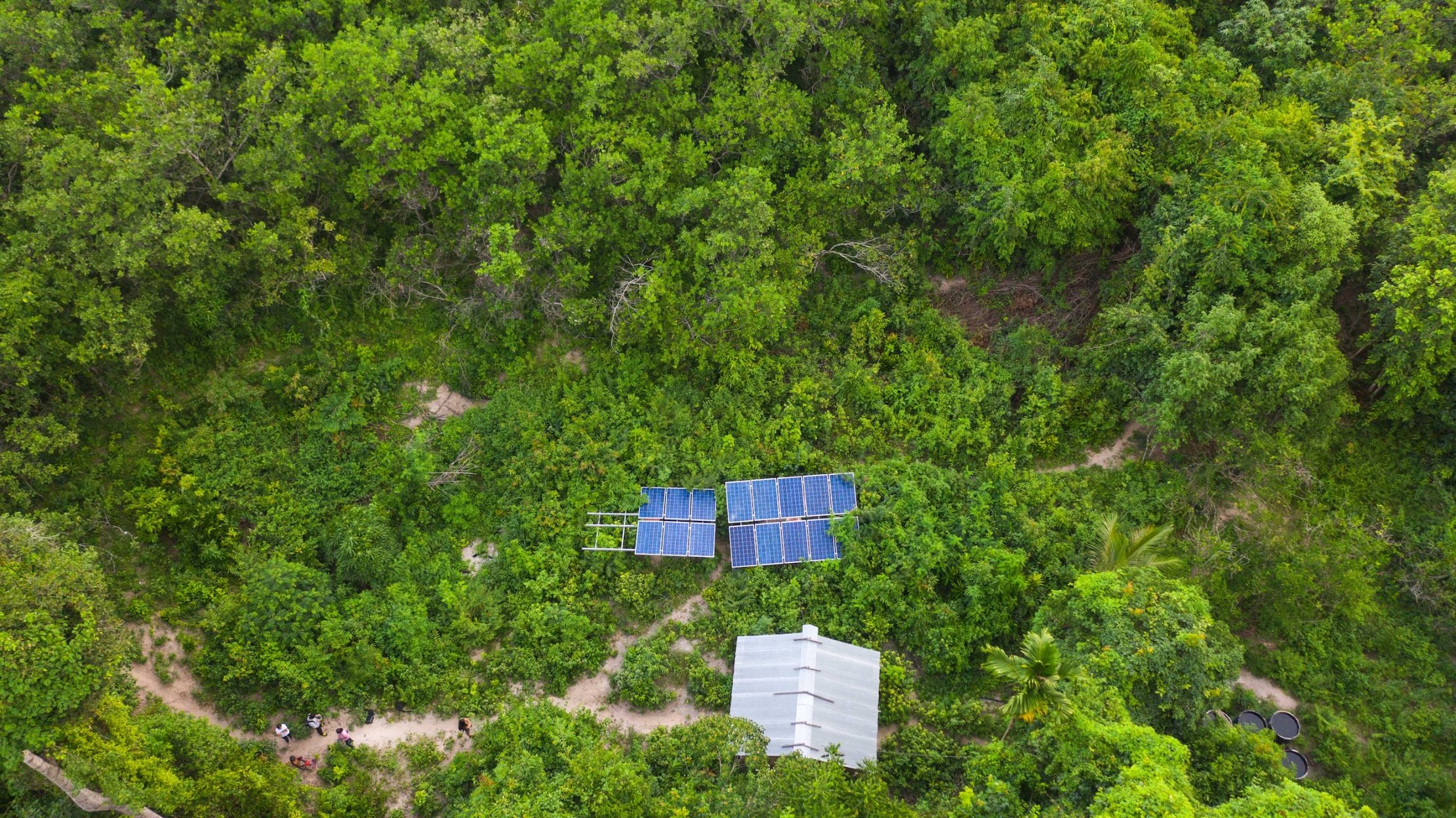 Aerial view of a small solar-panel installation and a metal-roofed structure surrounded by dense green forest.