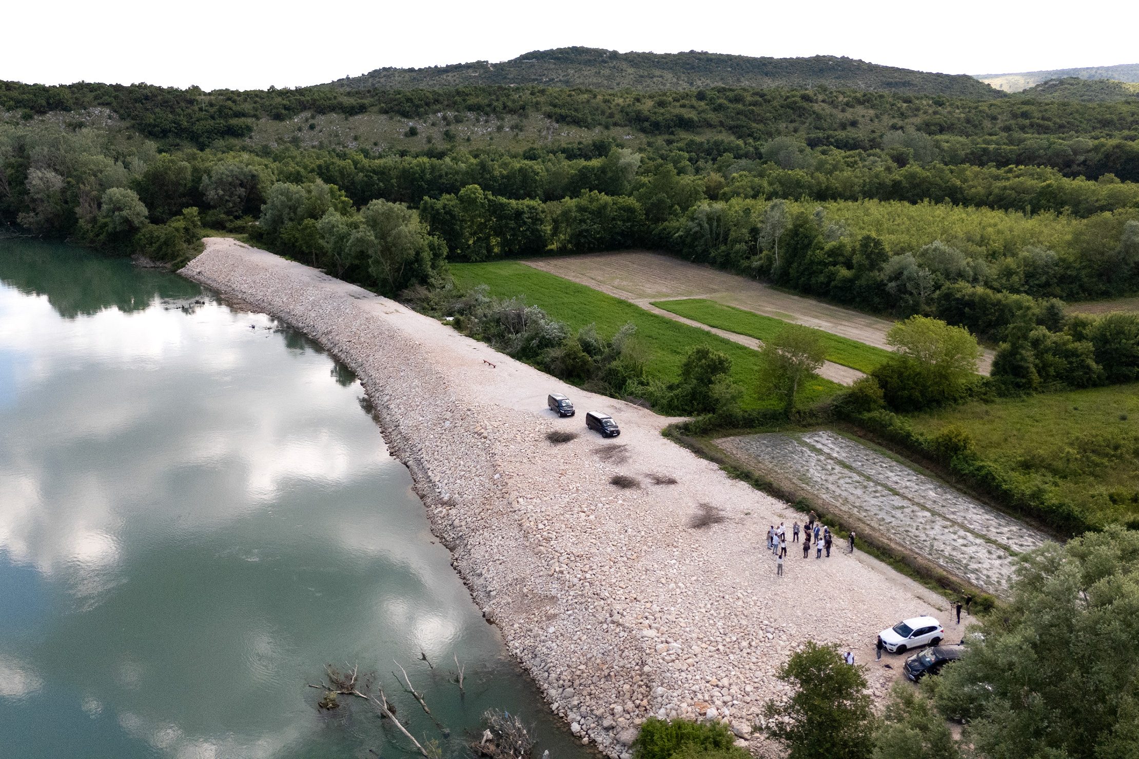 Aerial view of the restored Bojana/Buna River embankment in Montenegro, with cars and a small group of people gathered along the flood protection structure beside farmland and forest.