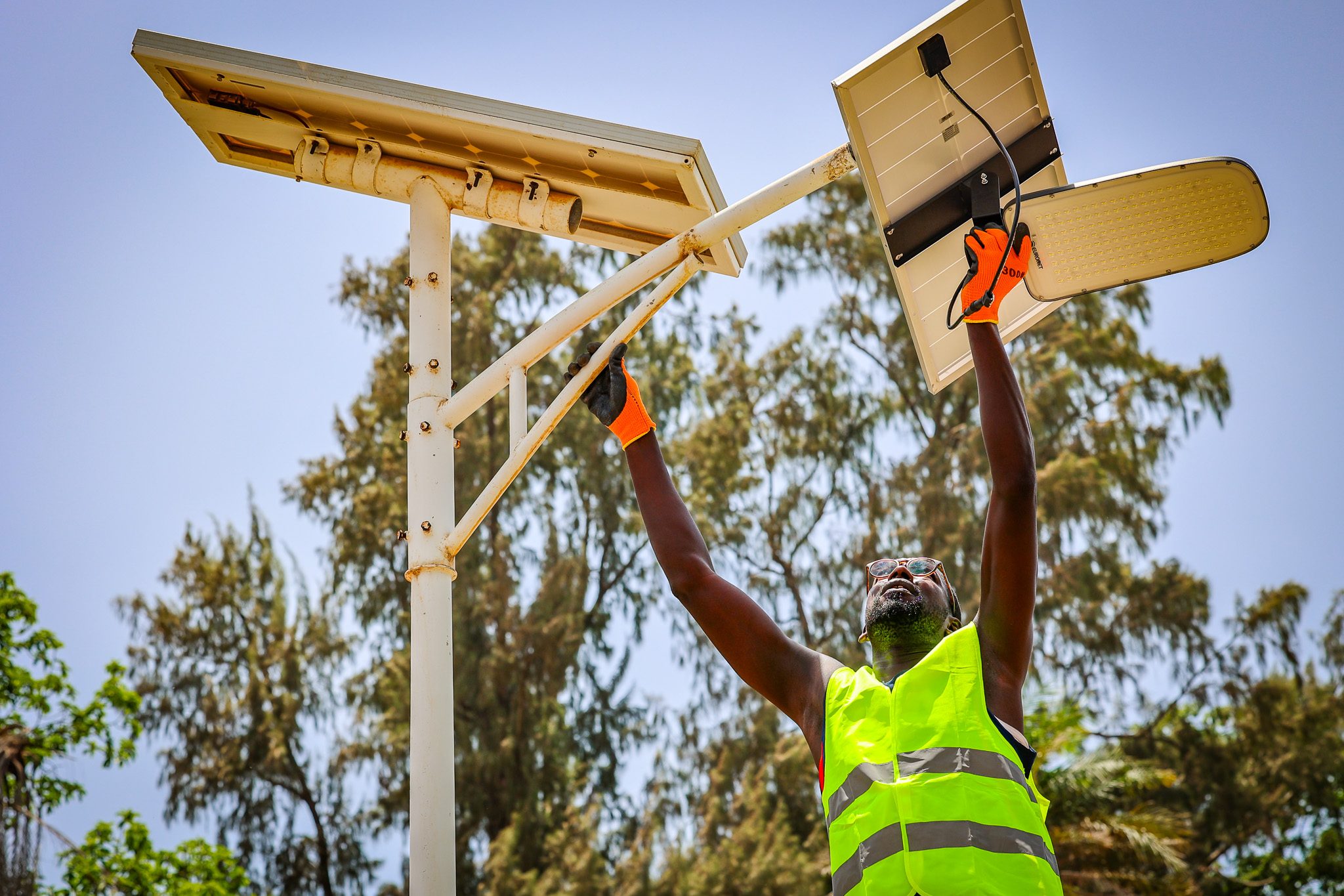 Photo: UNDP Senegal A technician in a high-visibility vest and orange gloves installing a solar-powered LED street light on a tall pole, with trees and a clear blue sky in the background in Casamance, Senegal
