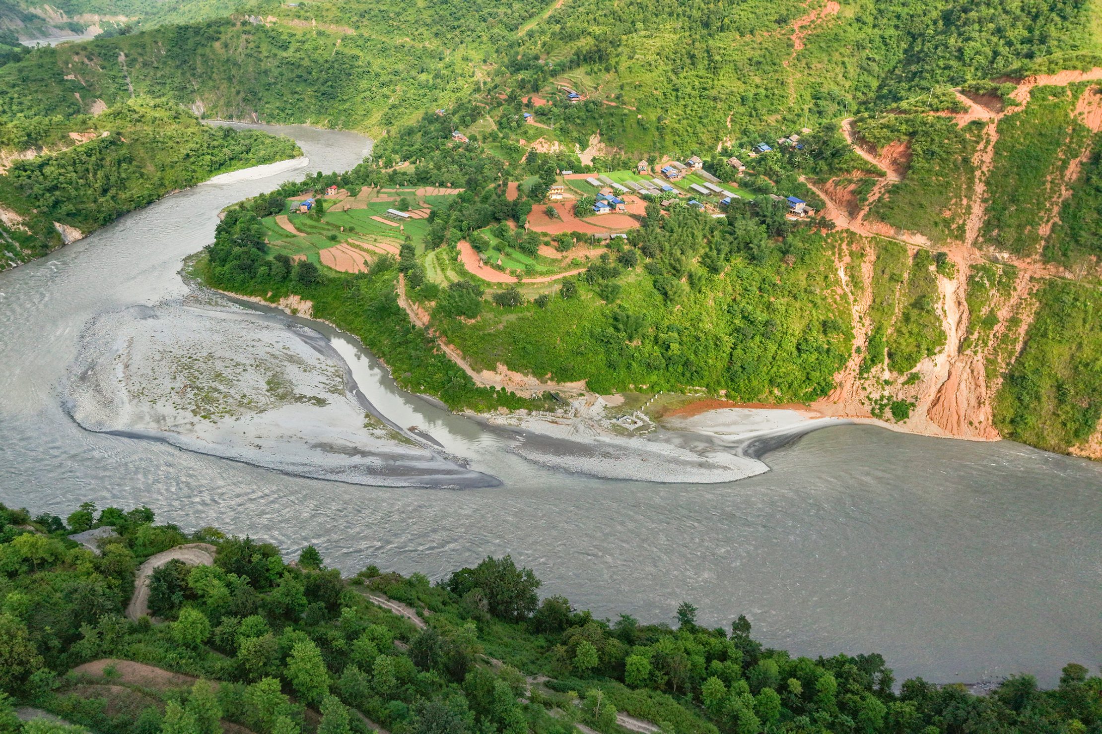 An aerial view of terraced farmland and homes along a winding river in Nepal’s hills, surrounded by green vegetation and steep slopes.
