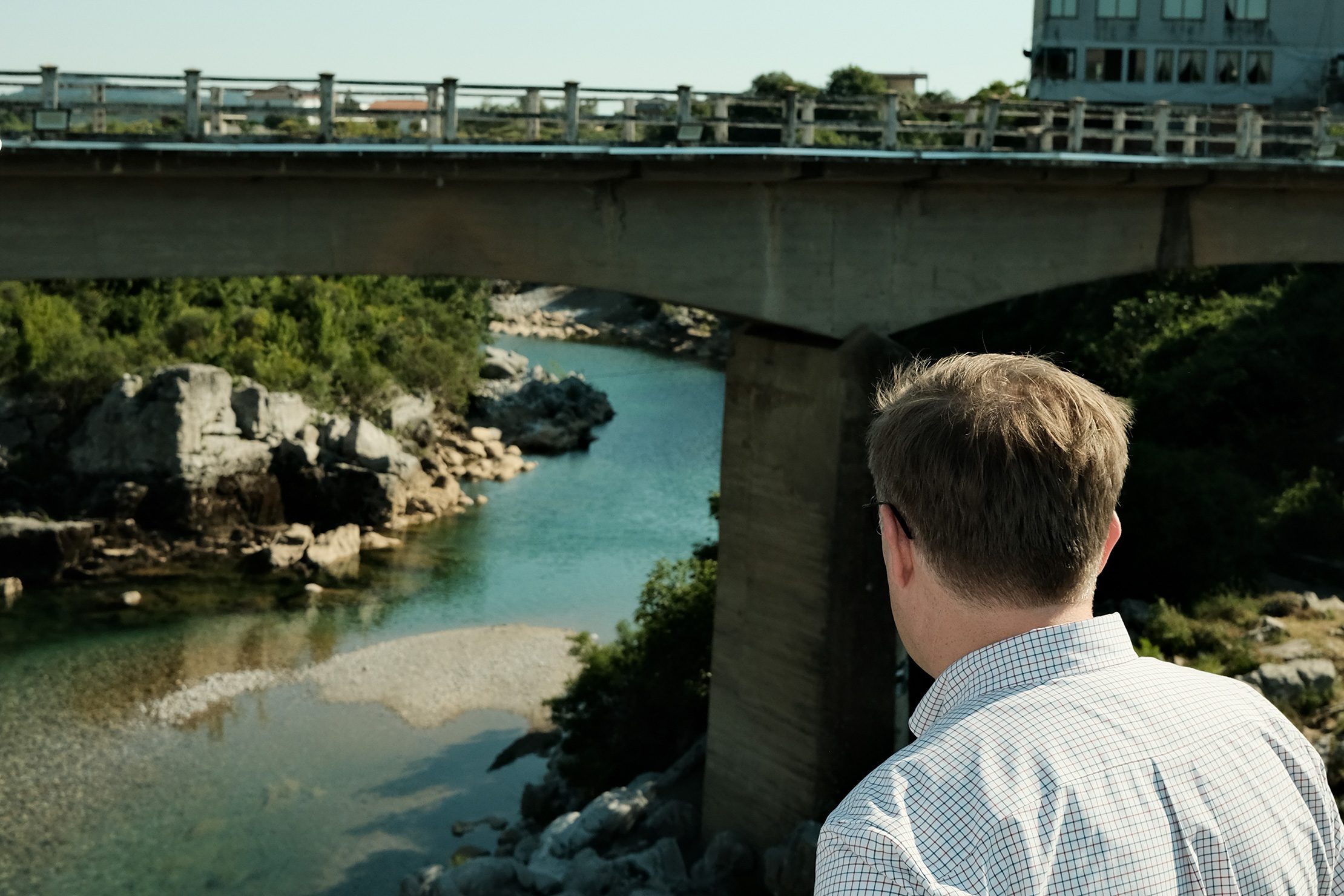 For residents of northern Albania, the dangers of flooding are all too familiar. Photo: Adaptation Fund A man in a checkered shirt looks out over a river flowing beneath a concrete bridge surrounded by rocks and greenery.