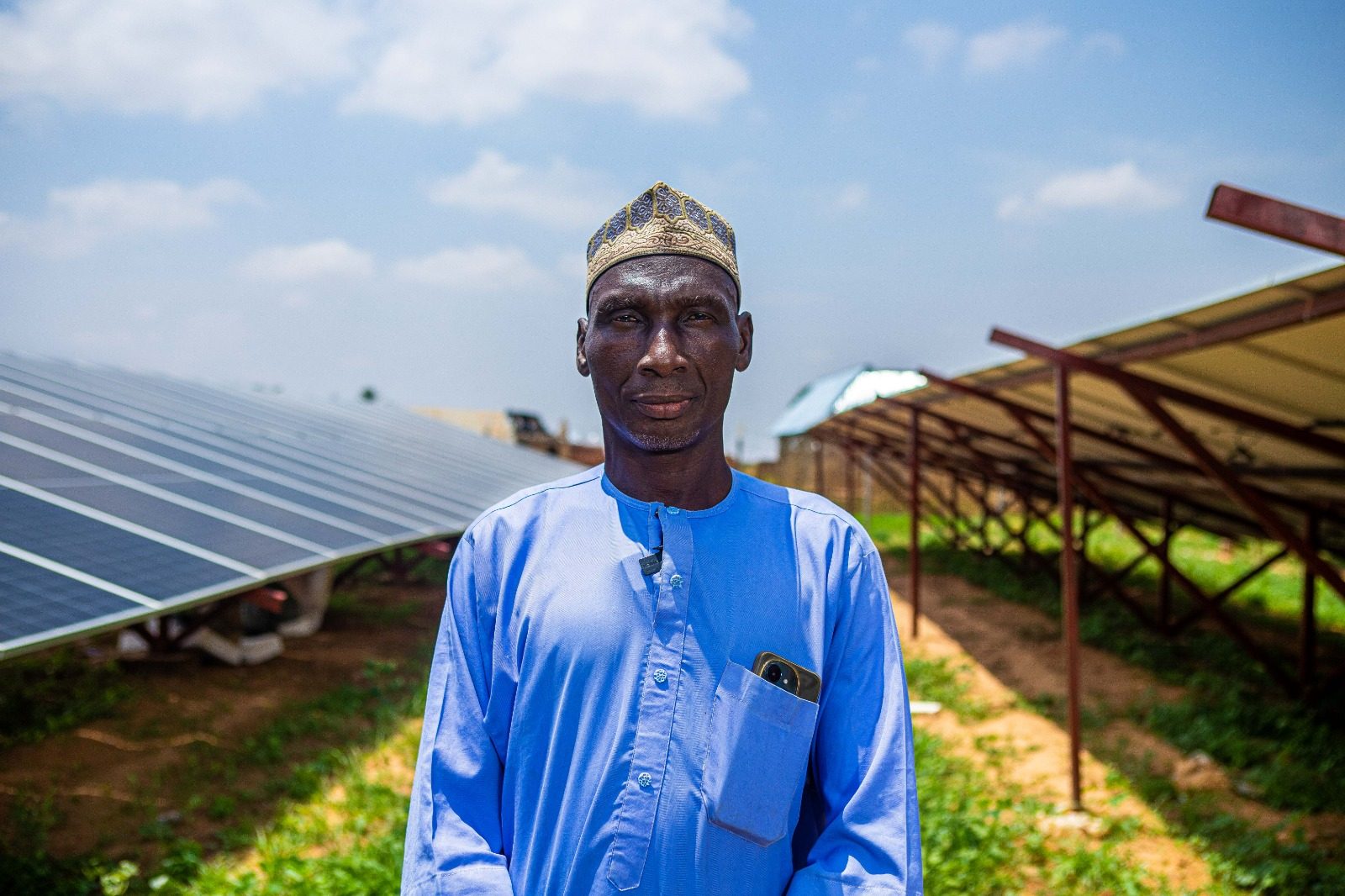 Man standing outdoors in front of solar panels, wearing a blue traditional outfit and a patterned cap under a bright sky.