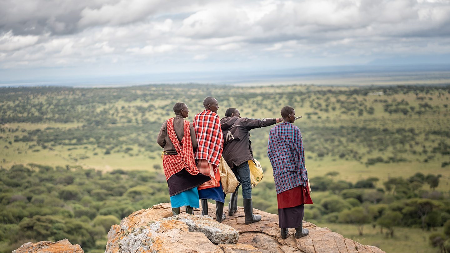 A group of Maasai men, dressed in traditional attire, stand on a rocky cliff in Loiborsiret, Tanzania, overlooking a vast landscape as one person points toward the horizon.