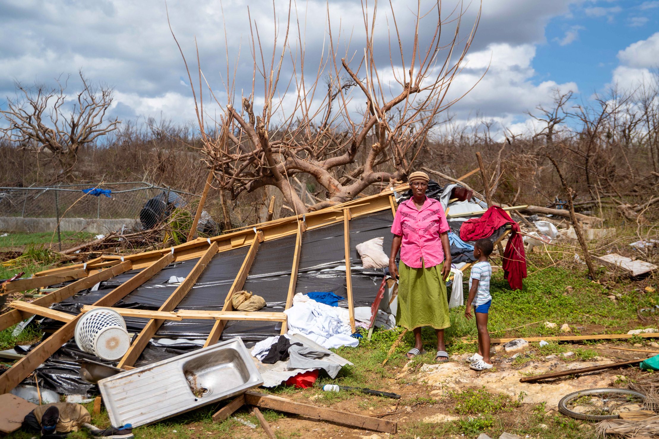 Woman and child stand beside a collapsed home and debris after Hurricane Melissa in Jamaica, showing the storm’s destruction to homes and livelihoods.