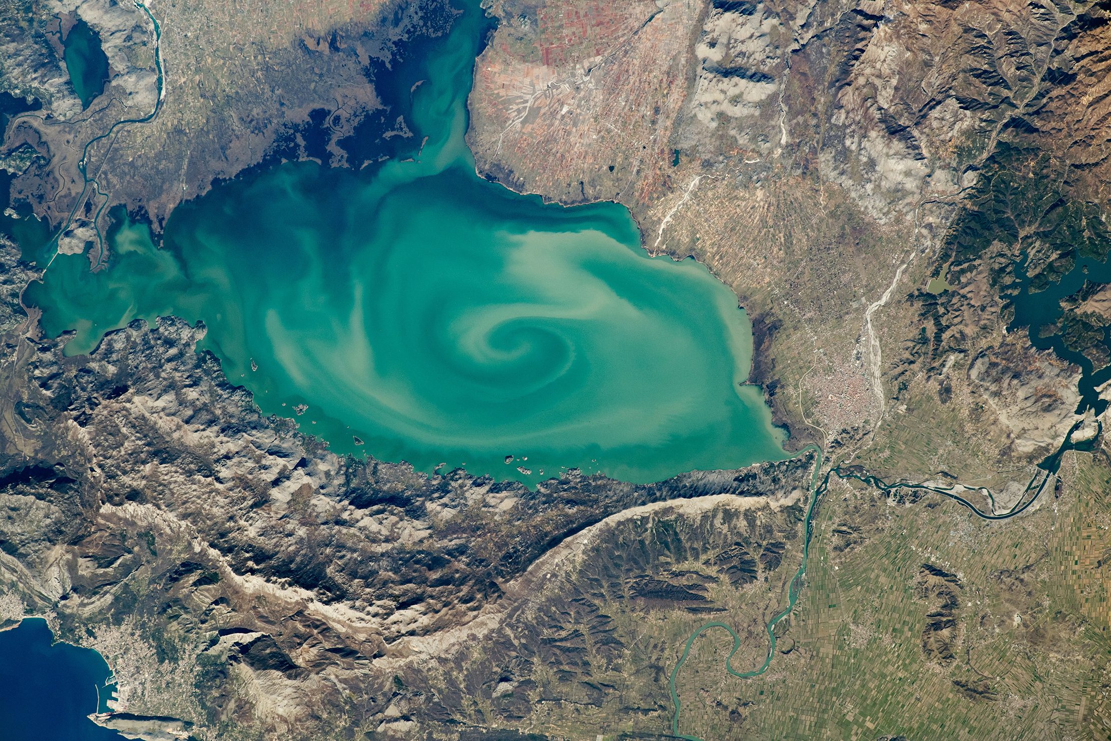 Satellite view of Lake Skadar on the border of Montenegro and Albania, part of the Drin River Basin. Image: NASA Satellite view of Lake Skadar on the border of Montenegro and Albania, showing turquoise waters with a swirling pattern surrounded by mountains, farmland, and river systems.