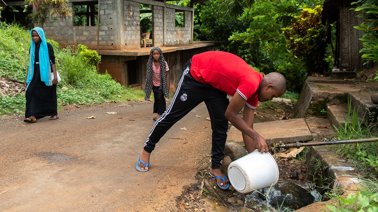 A young man fills a white bucket with water from a roadside stream while two women walk nearby in a rural area of Comoros, highlighting daily water access challenges.