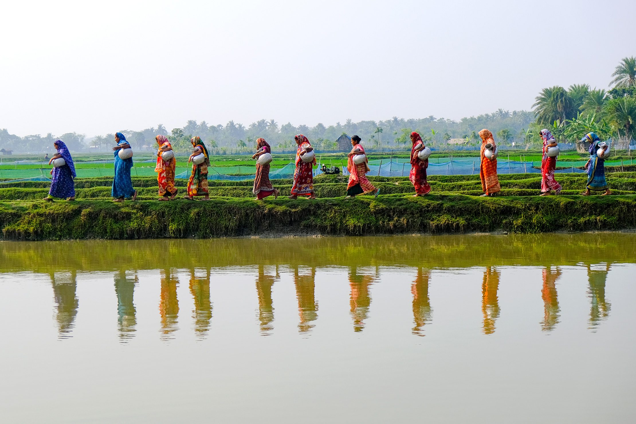 Women walk along an embankment in coastal Bangladesh carrying water pots, reflecting in nearby water.