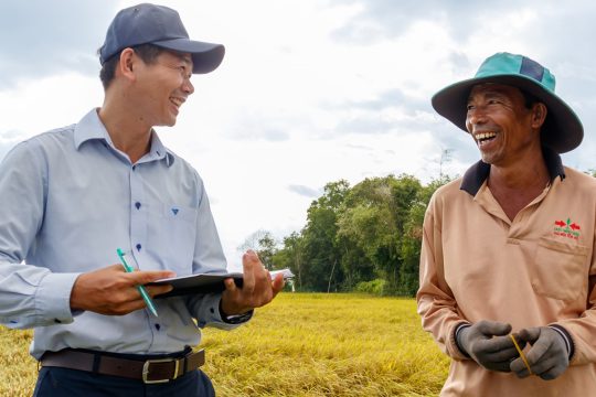 Un agricultor vietnamita sonriente y un funcionario del proyecto conversan en un campo de arroz, destacando los esfuerzos de adaptación climática impulsados ​​por la comunidad