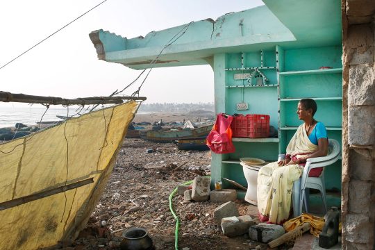 Woman sits inside a damaged coastal structure in Odisha, India, after cyclones and flooding, with debris and fishing boats visible along the shore.