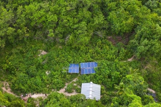 Aerial view of a small solar-panel installation and a metal-roofed structure surrounded by dense green forest.